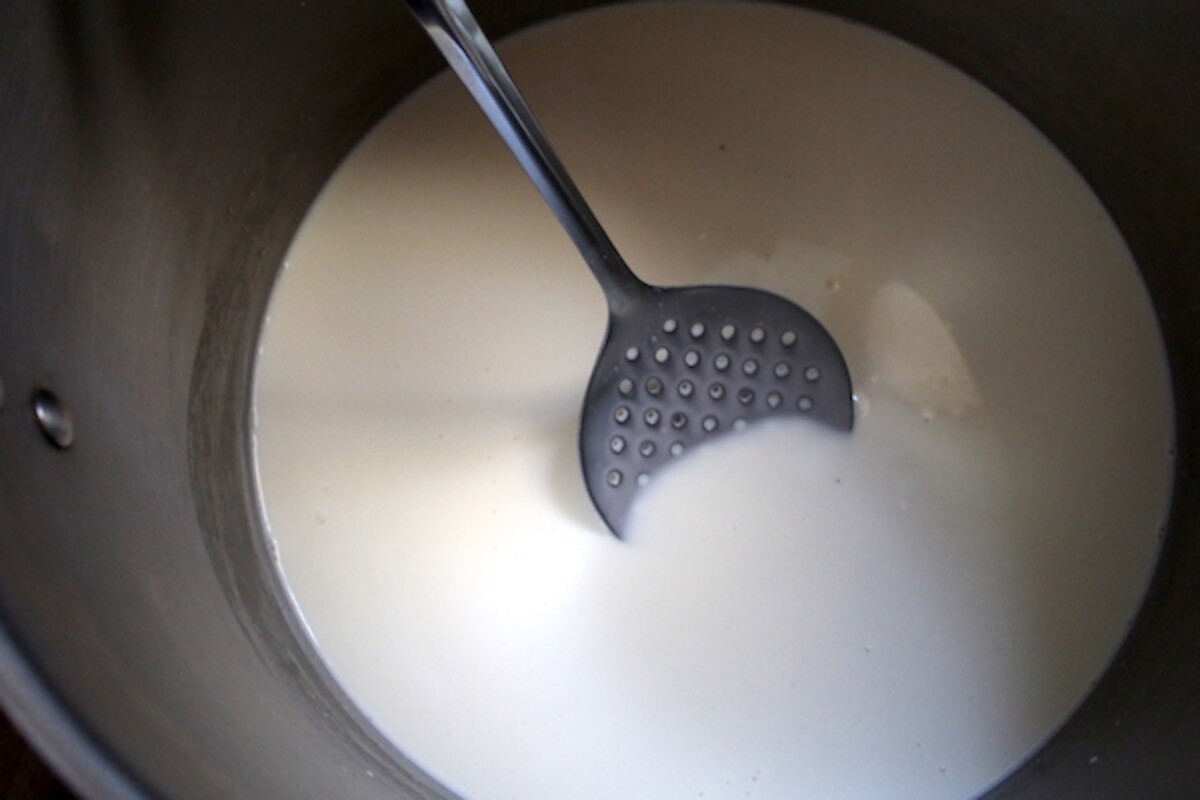Stirring the rennet into the raw milk with a large stainless steel slotted spoon.