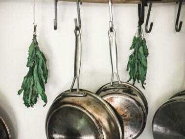Metal pots and pans and bundles of herbs hang from a stick on a kitchen wall.