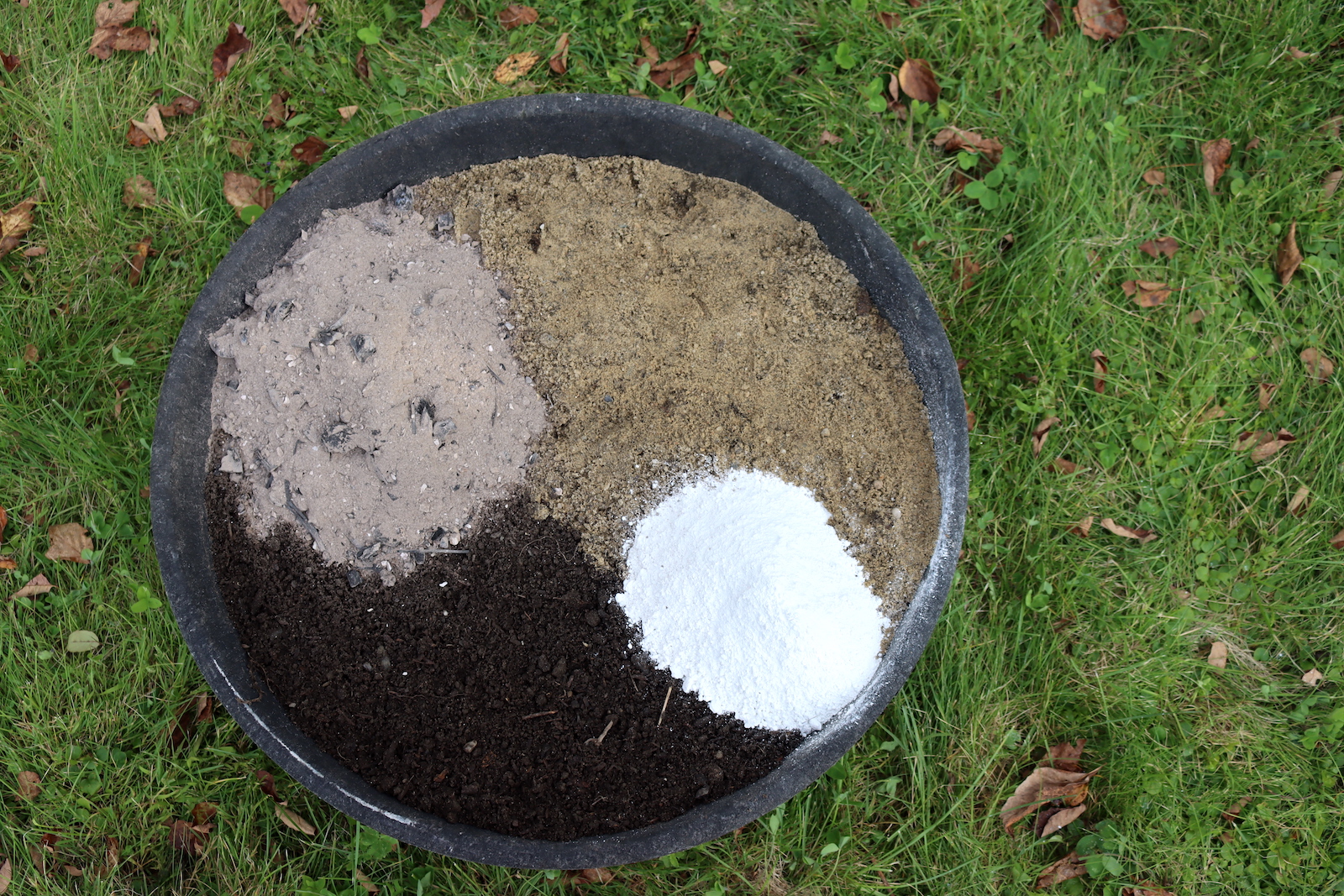 Separate piles of wood ash, sand, peat moss, and white powder in a black rubber pan, ready to mix into a chicken dust bath