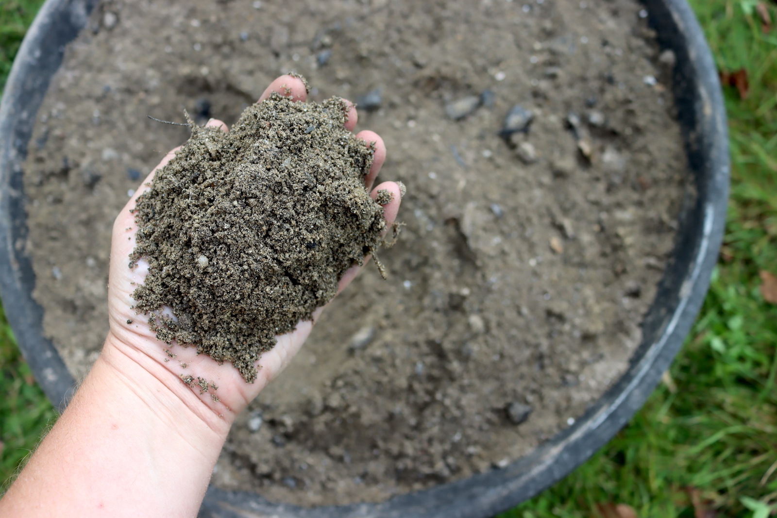Hand holding a scoop of mixed chicken dust bath over a rubber pan of sand, peat moss, and wood ash