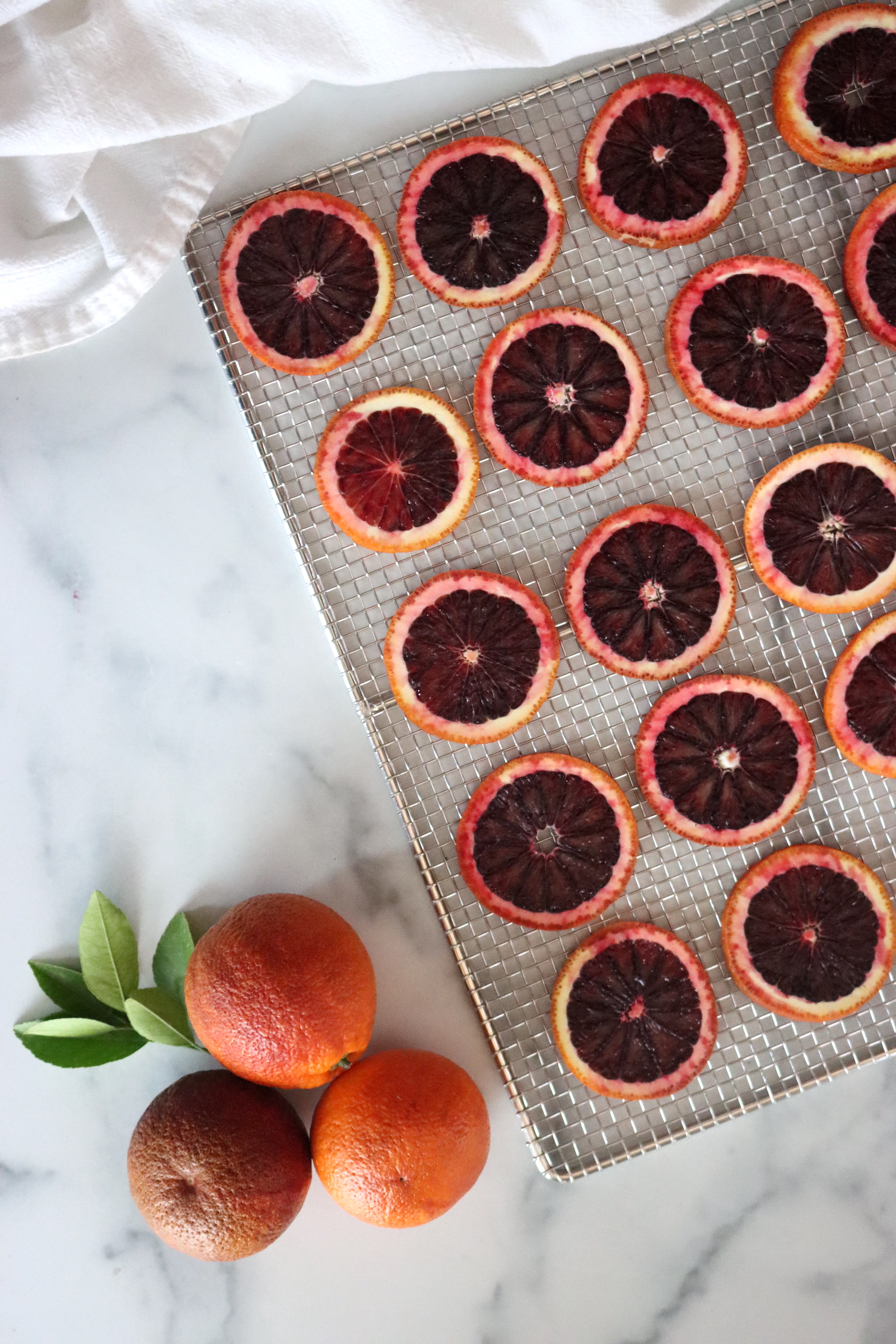 Dehydrated blood orange slices on a stainless mesh drying rack, with whole blood oranges and orange leaves