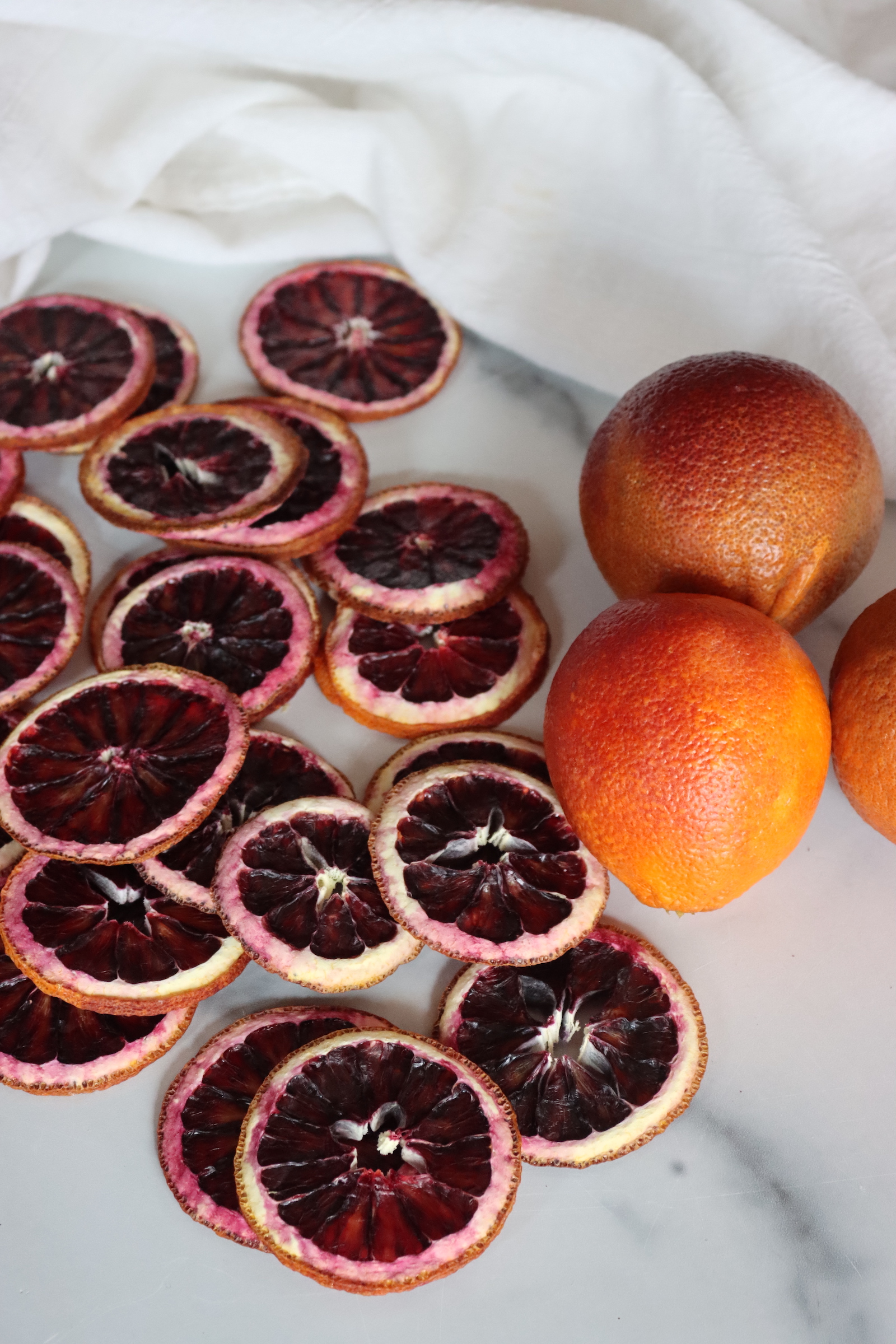 Dehydrated blood orange slices piled beside whole blood oranges on a white towel