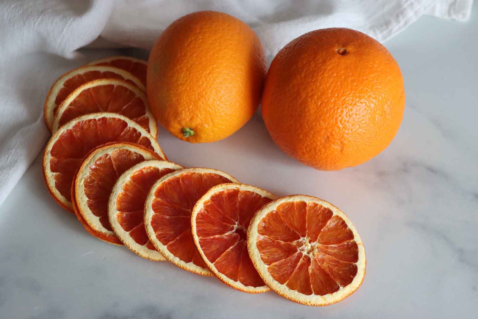 Dehydrated orange slices fanned out beside two whole oranges on a white marble counter
